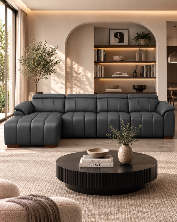 A modern dark gray tufted sectional sofa with a left-facing chaise lounge, styled in a warm, contemporary living room with a round black coffee table, beige rug, and built-in shelving in the background.