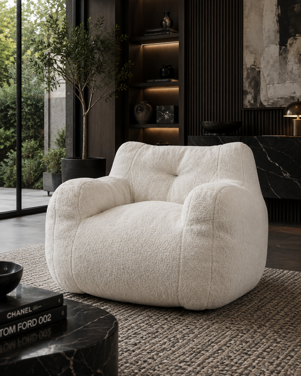 A plush, oversized boucle armchair in off-white is styled in a modern, dark-themed living room with a textured rug, a black marble side table, and a large window in the background.