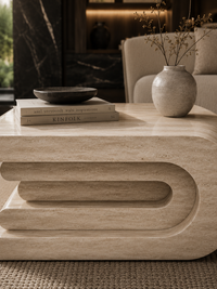 A modern curved stone coffee table in a warm beige travertine or marble finish, styled in a cozy living room setting with books, a small bowl, and a ceramic vase on top.