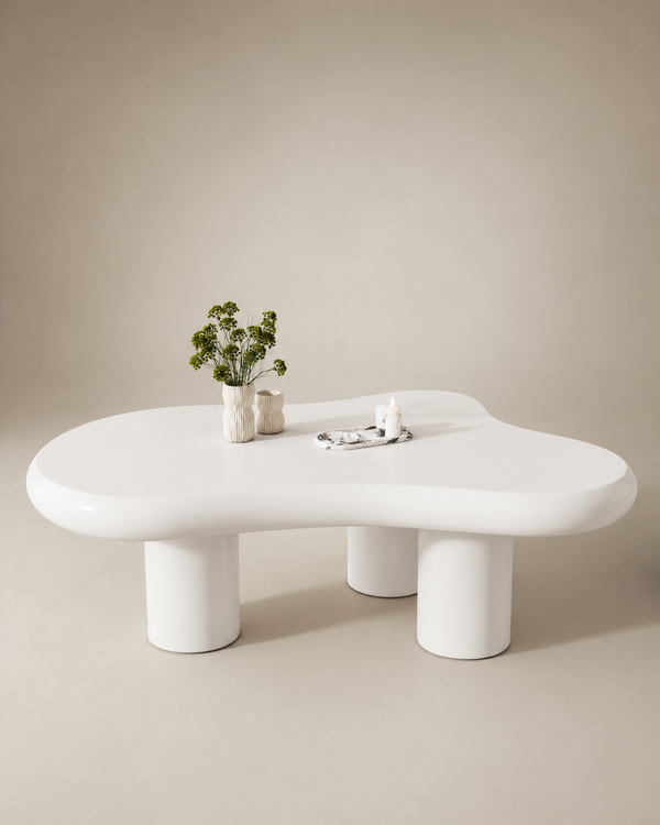 A glossy white, organic-shaped coffee table with thick cylindrical legs, styled with two small vases containing green stems and a small candle on a matching neutral background.