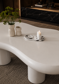A modern, glossy white wavy coffee table in a stylishly decorated living room, styled with two textured cream vases containing green plants and a small marble tray holding a lit candle.