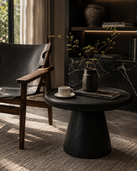 A dark round pedestal side table in a modern living room setting, styled with a small ceramic vase of branches, a book, and a cup and saucer. A dark leather and wood armchair is visible in the background next to a window.