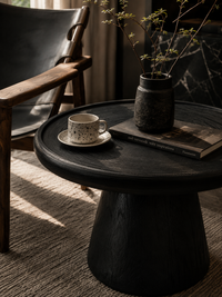 A round black wooden coffee table in a styled living room setting, holding a small speckled teacup on a saucer and a dark textured vase with dried branches, next to a wooden armchair.