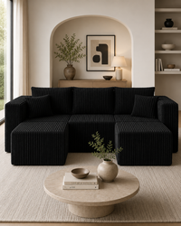 A modern sectional sofa in black ribbed fabric, styled in a minimalist living room with a round stone coffee table, neutral wall decor, and an arched alcove in the background.