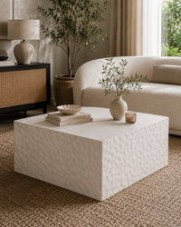 A modern square coffee table with a textured, hammered finish in a warm off-white or cream color, styled in a cozy living room setting with a beige sofa, woven rug, and potted plant.