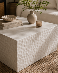 A modern textured coffee table in a warm cream tone, styled with a ceramic vase of greenery, a small amber glass candle holder, and a stack of books on a woven rug in a living room setting.