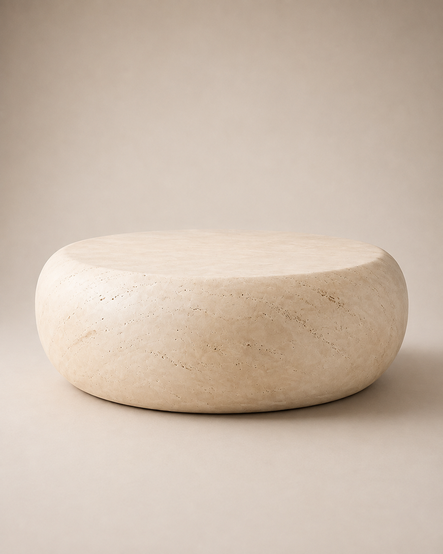 A round, low-profile stone ottoman or stool in a warm beige travertine-like material, photographed against a matching neutral beige studio background.