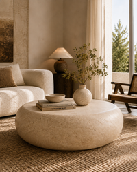 A round, low-profile coffee table made of textured beige travertine stone sits in a warm, modern living room. The table is styled with a small ceramic vase containing dried branches, a stack of books, and a small ceramic bowl.