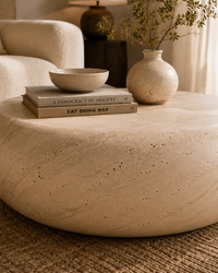 A round, low-profile stone ottoman or coffee table in a warm beige travertine-like finish, styled in a cozy living room setting with books, a small bowl, and a vase on top.