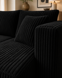 A close-up of a black corduroy sofa with matching throw pillow in a warm, modern living room setting.