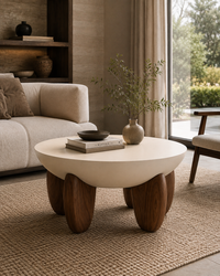 A modern round coffee table with a smooth off-white top and chunky dark brown wooden legs, styled in a minimalist living room next to a beige sofa and a large window.