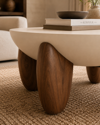 A modern low coffee table with a smooth cream-colored top and three sculptural dark wood legs, shown in a warm, neutral-toned living room setting on a woven rug.