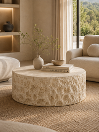 A round, textured stone coffee table in a warm beige tone, styled with a small vase and books in a modern, neutral-toned living room.