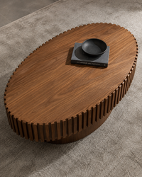 An oval wooden coffee table with a slatted, ribbed base and a rich medium-brown wood grain, styled on a textured gray rug with a black bowl on top.