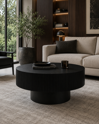 A round black ribbed coffee table in a modern living room setting with a beige sofa, textured rug, and dark wood shelving.