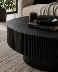 A round black coffee table with a vertically fluted, pleated side design, styled in a modern living room with a book, a small dark glass cup, and a black marble tray on top.