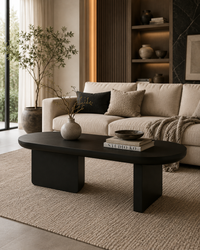 A modern minimalist living room featuring a dark oval coffee table with blocky legs, styled on a textured beige rug in front of a cream-colored sofa with neutral throw pillows.