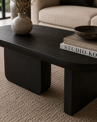 A modern black coffee table with a rounded top and blocky legs, styled in a living room setting with a textured beige rug, a gray ceramic vase, and decorative books.