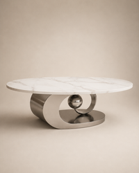 A modern round coffee table with a white marble top and a sculptural brushed metal base featuring a central polished sphere, set against a warm beige background.