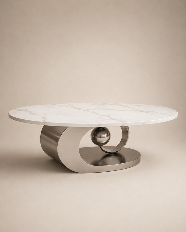 A modern round coffee table with a white marble top and a sculptural brushed metal base featuring a central polished sphere, set against a warm beige background.