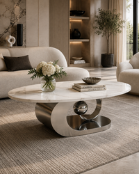 A modern oval coffee table with a white marble top and a sculptural polished metal base, styled in a contemporary living room with a cream sofa, textured rug, and decorative flowers.