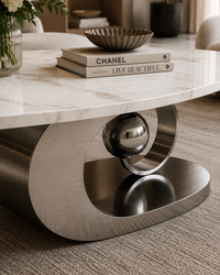 A modern round coffee table with a white marble top and a brushed stainless steel sculptural base featuring a central metallic sphere, shown in a styled living room setting.