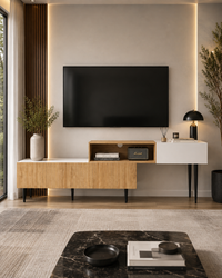 A modern living room setup featuring a wall-mounted black TV above a low wooden media console with a white floating side cabinet and black legs. The room has warm ambient lighting, a textured beige rug, a black marble coffee table in the foreground, and decorative plants.