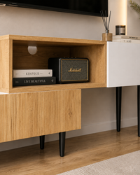 A modern wooden media console with an open shelf holding a black Marshall speaker and books, next to a white cabinet panel on a light wood floor.