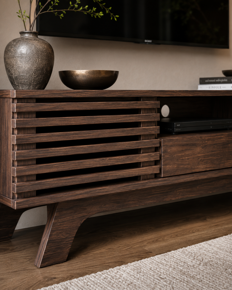 A dark brown wooden TV stand or media console with a slatted cabinet door and angled legs, shown in a styled living room setting with a vase, bowl, and wall-mounted TV in the background.