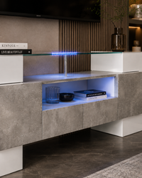 Modern floating TV console with a textured gray stone-look front, white geometric side blocks, a glass shelf, and integrated blue LED lighting, shown in a styled interior setting.
