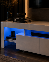 A modern white glossy media console in a living room setting, illuminated by blue LED under-shelf lighting. The console features open shelving on the left with decorative books and a vase, and closed cabinet doors on the right.