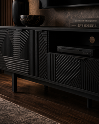 A modern black TV stand or media console with geometric textured paneling, shown in a styled living room setting with dark wood flooring, a rug, and decorative objects on top.