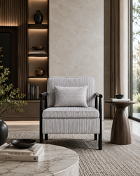 A modern upholstered armchair with a striped gray and white fabric, black wooden arms and legs, and a matching striped throw pillow, set in a contemporary living room next to a wooden side table.