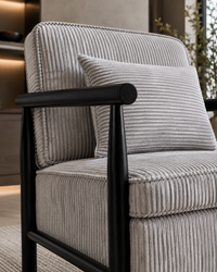A close-up of a modern armchair with ribbed light gray corduroy upholstery and a matching throw pillow, set against a softly blurred interior background.