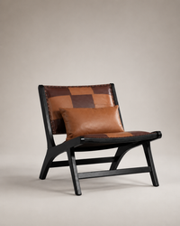 A modern lounge chair with a black wooden frame and a brown patchwork leather seat and backrest, accompanied by a matching brown leather lumbar pillow, set against a neutral beige background.