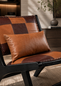 A close-up of a modern armchair with a patchwork leather backrest and seat cushion, styled with a matching brown leather lumbar pillow in a warm, sunlit interior setting.