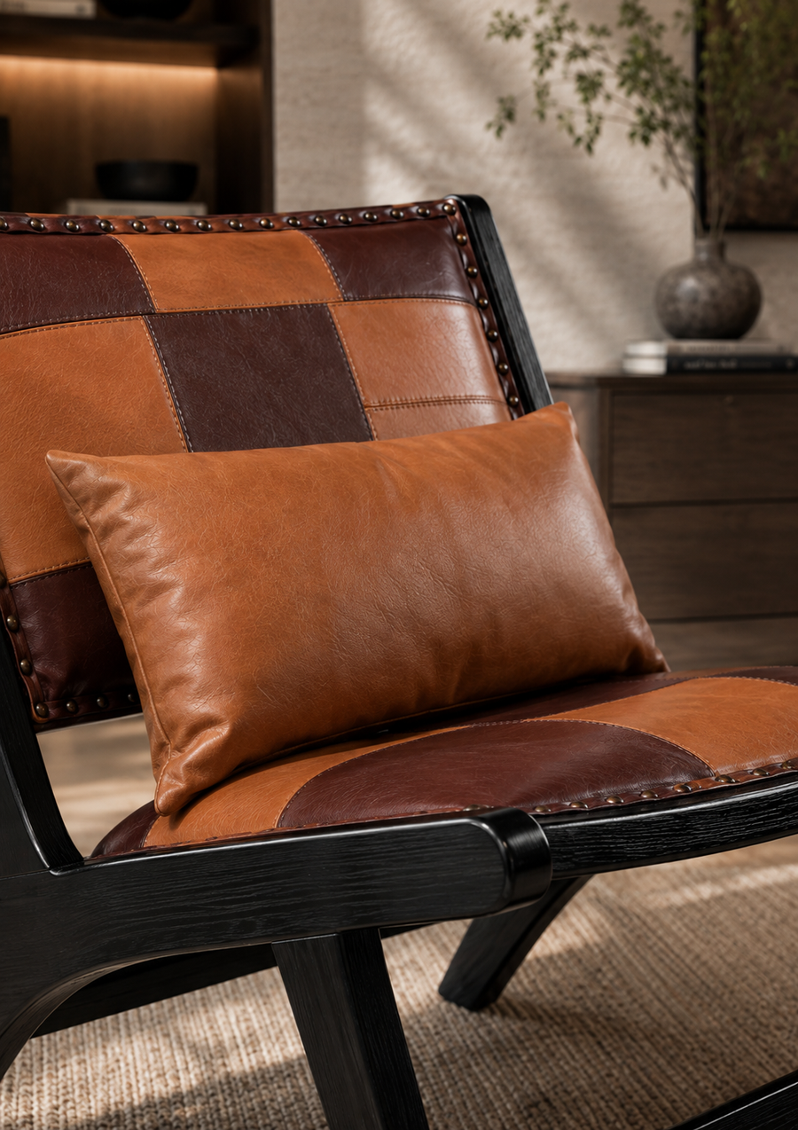 A close-up of a modern armchair with a patchwork leather backrest and seat cushion, styled with a matching brown leather lumbar pillow in a warm, sunlit interior setting.