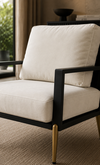 A modern accent chair with cream-colored cushions and a black metal frame, featuring tapered gold legs, shown in a styled interior setting.