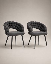Two modern dark gray upholstered dining chairs with sculptural rounded armrests and tapered black legs, shown on a plain warm beige studio background.