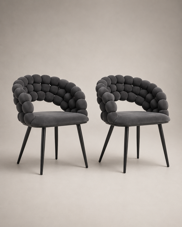 Two modern dark gray upholstered dining chairs with sculptural rounded armrests and tapered black legs, shown on a plain warm beige studio background.