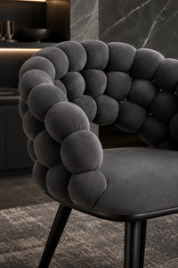 A close-up of a modern upholstered accent chair with a sculptural, bubble-tufted backrest in dark charcoal gray velvet, set in a contemporary interior with dark stone walls and flooring.