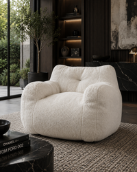 A plush, oversized boucle armchair in off-white is styled in a modern, dark-themed living room with a textured rug, a black marble side table, and a large window in the background.