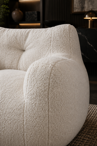 Close-up of a plush, cream-colored bouclé armchair in a modern, dark-toned living room setting.
