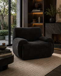 A dark charcoal gray plush armchair with rounded arms, positioned on a textured beige rug in a modern, dimly lit living room with a fireplace and bookshelves.