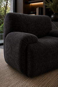A close-up of a modern dark charcoal gray bouclé armchair placed on a woven beige rug in a dimly lit interior setting.