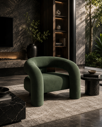 A modern curved armchair upholstered in deep olive green velvet, styled in a luxurious, dark-toned living room with a textured rug, marble side tables, and a dark wood shelving unit.