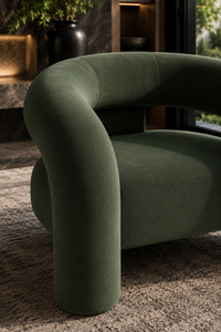 A modern curved armchair upholstered in dark olive green velvet, shown in a stylishly decorated interior setting with a textured rug and warm ambient lighting.