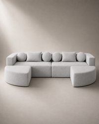 A light gray modular sectional sofa with two ottomans, styled in a minimalist studio setting with a matching beige background.