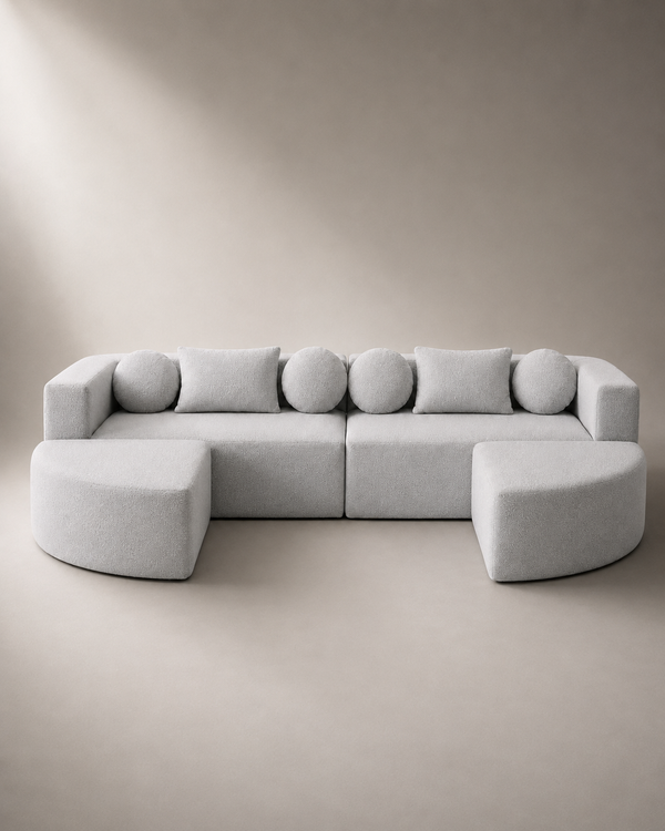A light gray modular sectional sofa with two ottomans, styled in a minimalist studio setting with a matching beige background.