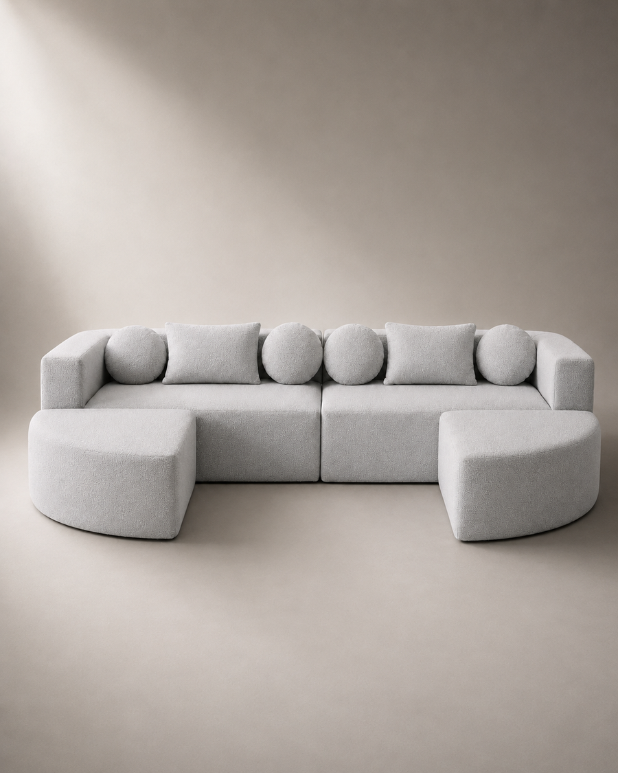 A light gray modular sectional sofa with two ottomans, styled in a minimalist studio setting with a matching beige background.